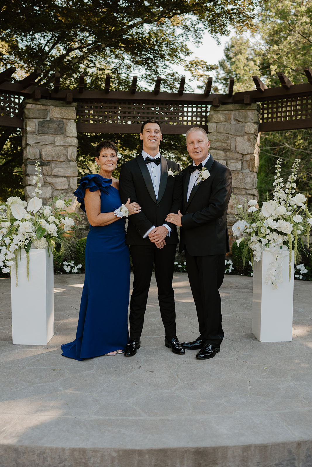 Groom posing for a portrait with his parents under a wooden arbor decorated with white flowers.